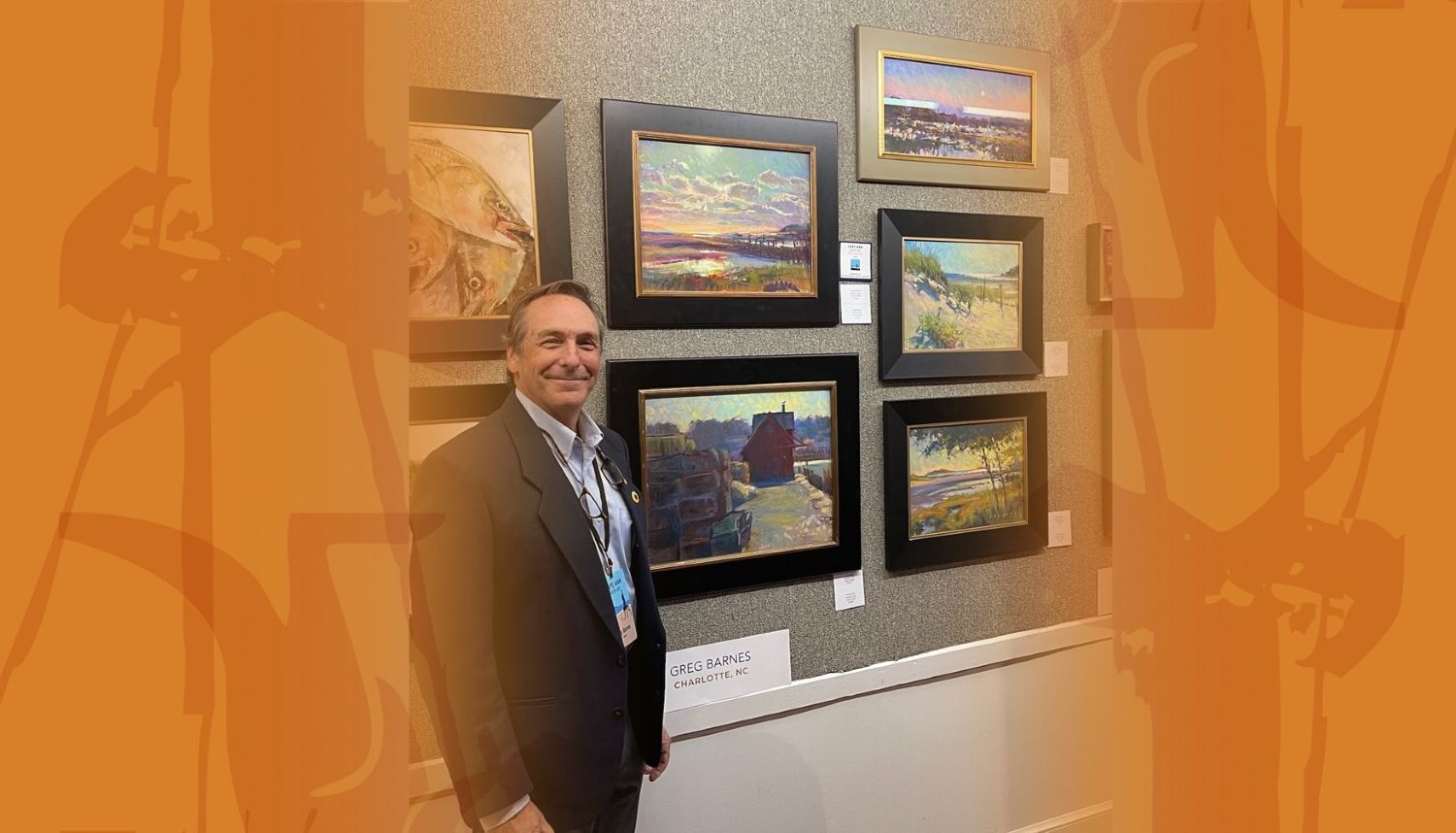 A man wearing a suit stands in front of a wall of landscape paintings, smiling